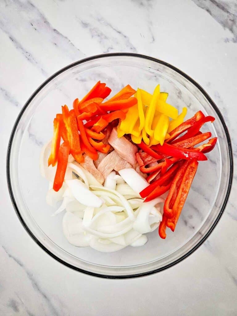 sliced chicken breast, bell peppers and onions in a glass bowl