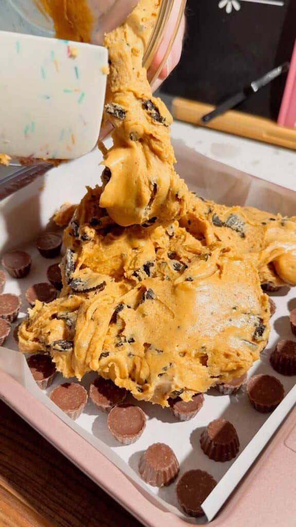 peanut butter oreo fudge mixture being poured over mini reese's peanut butter cups in a parchment lined baking pan