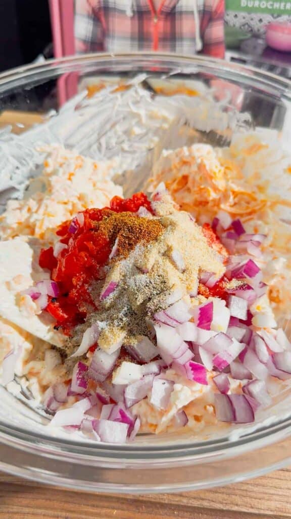spices, diced pimentos and diced red onions on top of cream cheese, mayo and cheddar mixture in a mixing bowl
