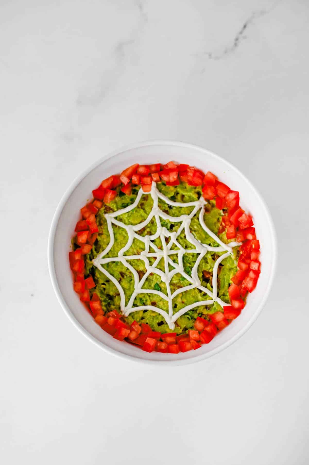 This Halloween Guacamole is the ultimate spooky appetizer for any spooky celebration! Creamy homemade guacamole with avocado, Roma tomatoes, red onion, fresh cilantro, and lime juice gets a festive makeover with an easy sour cream spider web on top. This Spider Web Guacamole is a fun Halloween dip that's ready in just 15 minutes and perfect with tortilla chips, crackers, or fresh veggies. If you need Easy Halloween Appetizers, Halloween Party Food, Halloween Dip Recipes, Spooky Halloween Food, Halloween Snacks for Kids, or Halloween Guacamole Ideas, this simple recipe belongs on your menu. Add diced tomatoes around the edge and a plastic spider for an extra creepy finish. It's an easy Halloween party appetizer that's fresh, colorful, delicious, and perfect for a Halloween snack table!