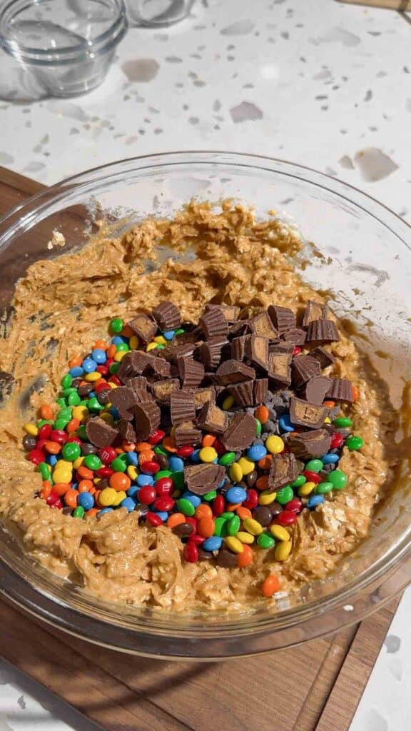 chopped peanut butter cups, mini M&Ms and mini chocolate chips on top of peanut butter mixture in a glass bowl