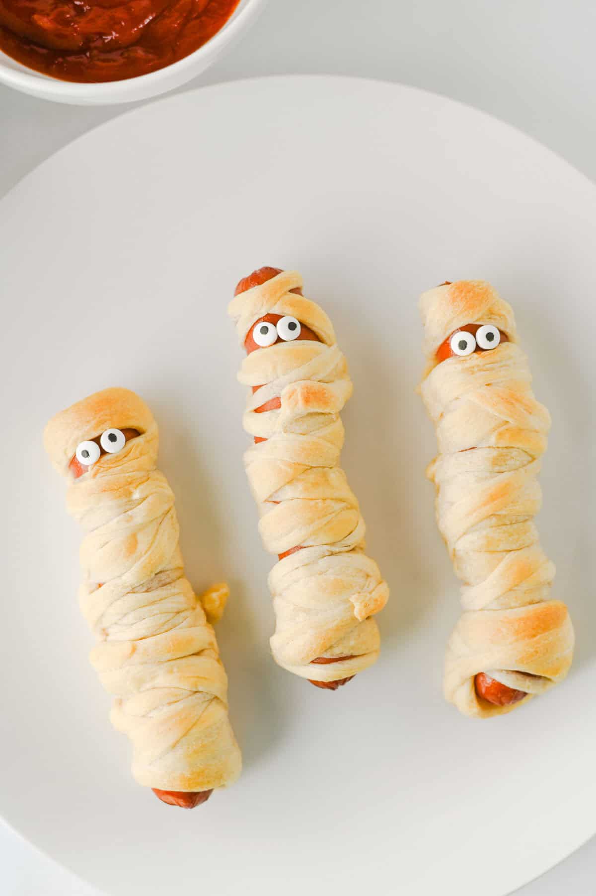 These Mummy Hot Dogs are the perfect easy Halloween dinner, spooky party snack, or kid-friendly Halloween appetizer! Made with hot dogs wrapped in flaky crescent roll dough, these adorable Crescent Roll Mummy Hot Dogs are quick to prep and baked until golden. Add candy eyes for a fun finishing touch, then serve with ketchup and mustard for dipping. If you need Easy Halloween Food Ideas, Halloween Party Food For Kids, Halloween Hot Dog Ideas, Halloween Finger Foods, Spooky Halloween Snacks, or Halloween Dinner Ideas For Kids, this simple recipe is a must-try. These Halloween Mummy Dogs are great for Halloween parties, movie nights, or a festive family dinner. Save this Mummy Hot Dogs Recipe for an easy Halloween snack that's cute, delicious, and made with a few simple ingredients!