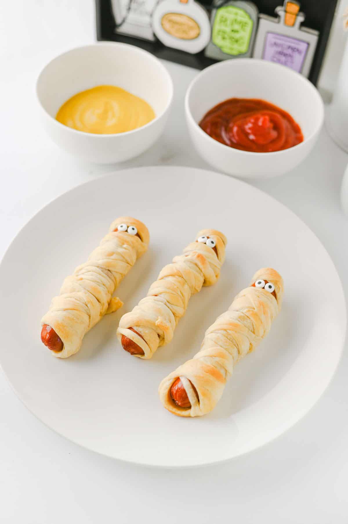 These Mummy Hot Dogs are the perfect easy Halloween dinner, spooky party snack, or kid-friendly Halloween appetizer! Made with hot dogs wrapped in flaky crescent roll dough, these adorable Crescent Roll Mummy Hot Dogs are quick to prep and baked until golden. Add candy eyes for a fun finishing touch, then serve with ketchup and mustard for dipping. If you need Easy Halloween Food Ideas, Halloween Party Food For Kids, Halloween Hot Dog Ideas, Halloween Finger Foods, Spooky Halloween Snacks, or Halloween Dinner Ideas For Kids, this simple recipe is a must-try. These Halloween Mummy Dogs are great for Halloween parties, movie nights, or a festive family dinner. Save this Mummy Hot Dogs Recipe for an easy Halloween snack that's cute, delicious, and made with a few simple ingredients!