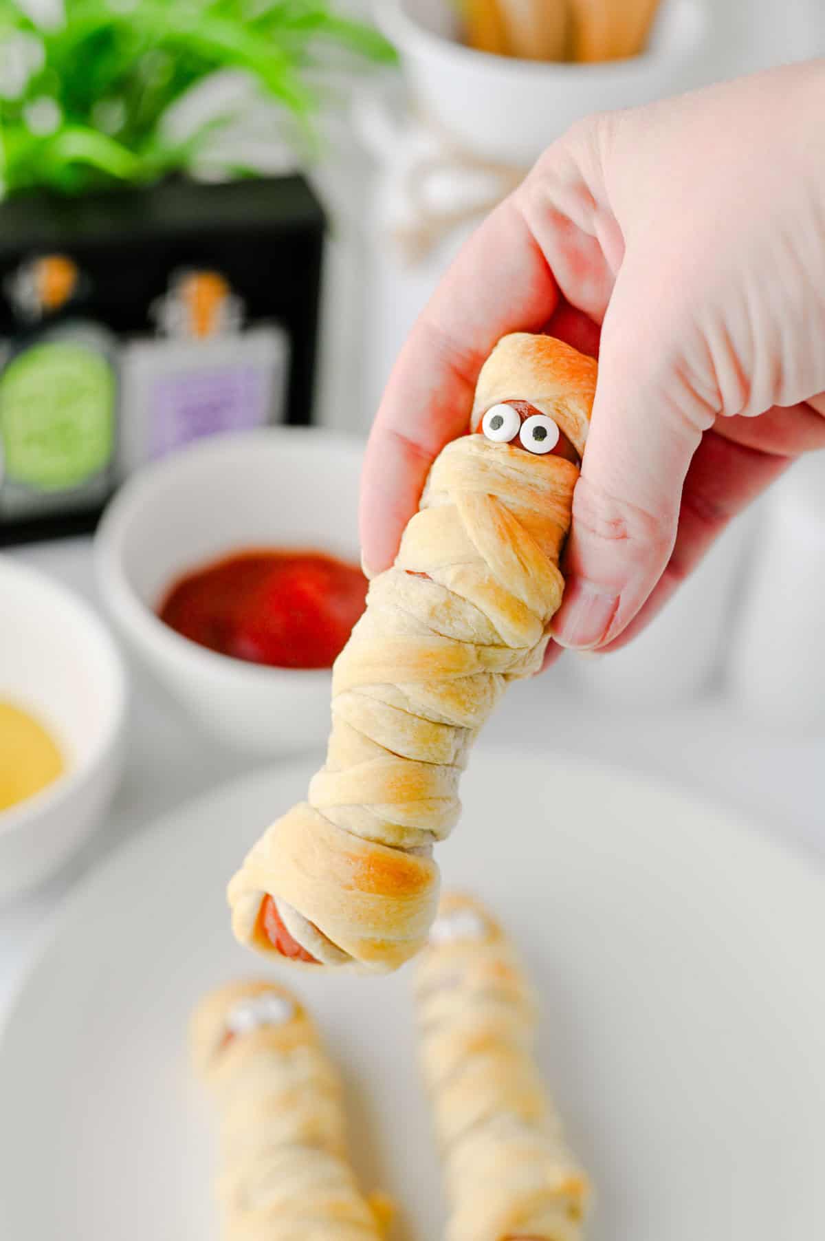 These Mummy Hot Dogs are the perfect easy Halloween dinner, spooky party snack, or kid-friendly Halloween appetizer! Made with hot dogs wrapped in flaky crescent roll dough, these adorable Crescent Roll Mummy Hot Dogs are quick to prep and baked until golden. Add candy eyes for a fun finishing touch, then serve with ketchup and mustard for dipping. If you need Easy Halloween Food Ideas, Halloween Party Food For Kids, Halloween Hot Dog Ideas, Halloween Finger Foods, Spooky Halloween Snacks, or Halloween Dinner Ideas For Kids, this simple recipe is a must-try. These Halloween Mummy Dogs are great for Halloween parties, movie nights, or a festive family dinner. Save this Mummy Hot Dogs Recipe for an easy Halloween snack that's cute, delicious, and made with a few simple ingredients!