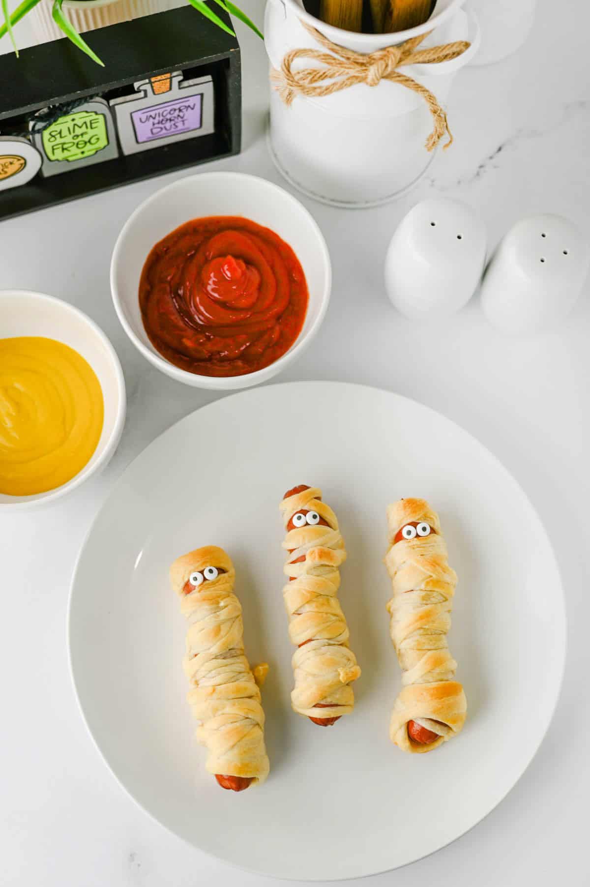 These Mummy Hot Dogs are the perfect easy Halloween dinner, spooky party snack, or kid-friendly Halloween appetizer! Made with hot dogs wrapped in flaky crescent roll dough, these adorable Crescent Roll Mummy Hot Dogs are quick to prep and baked until golden. Add candy eyes for a fun finishing touch, then serve with ketchup and mustard for dipping. If you need Easy Halloween Food Ideas, Halloween Party Food For Kids, Halloween Hot Dog Ideas, Halloween Finger Foods, Spooky Halloween Snacks, or Halloween Dinner Ideas For Kids, this simple recipe is a must-try. These Halloween Mummy Dogs are great for Halloween parties, movie nights, or a festive family dinner. Save this Mummy Hot Dogs Recipe for an easy Halloween snack that's cute, delicious, and made with a few simple ingredients!