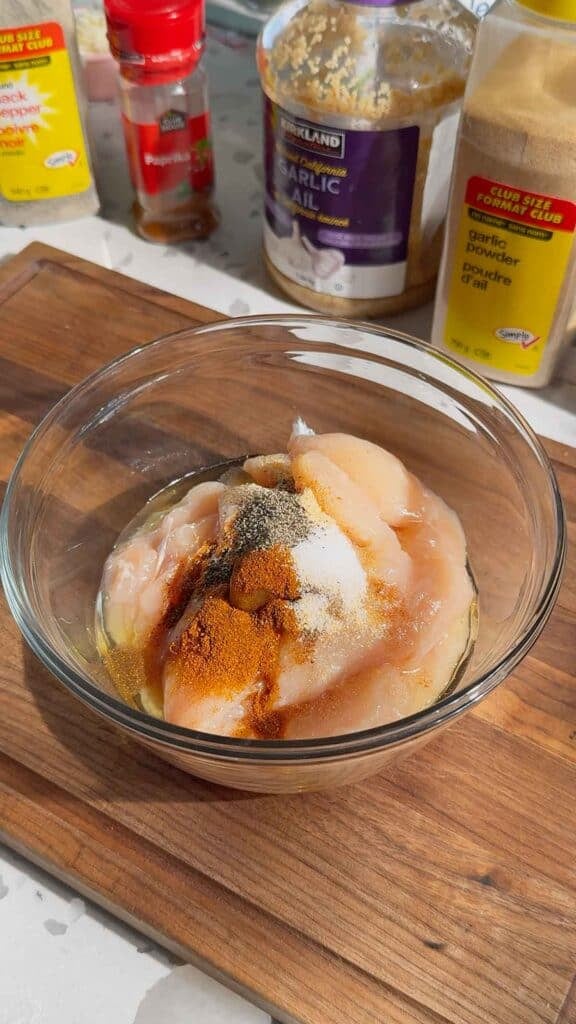 seasonings on top of raw chicken breast slices in a glass bowl