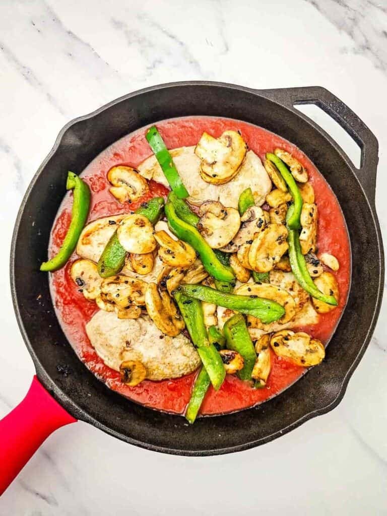 sliced green peppers and mushrooms on top of chicken breasts in a skillet with tomato sauce