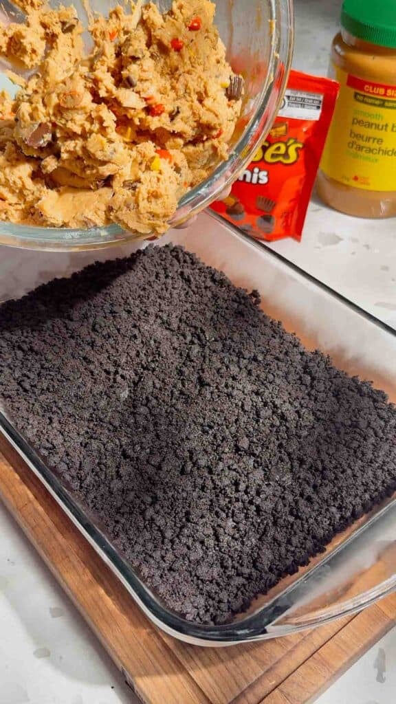 pouring peanut butter cheesecake mixture over oreo crust in a baking dish.