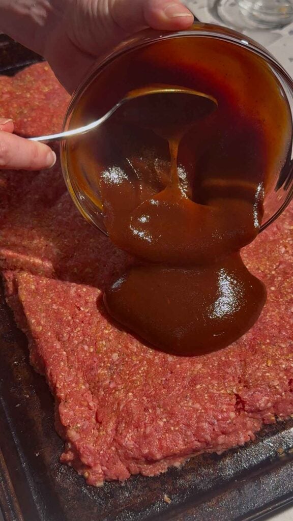 pouring bbq sauce over raw meatloaf on a sheet pan