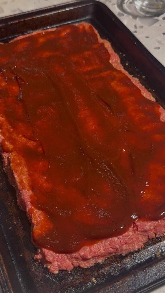bbq sauce mixture spread on raw meatloaf on a sheet pan