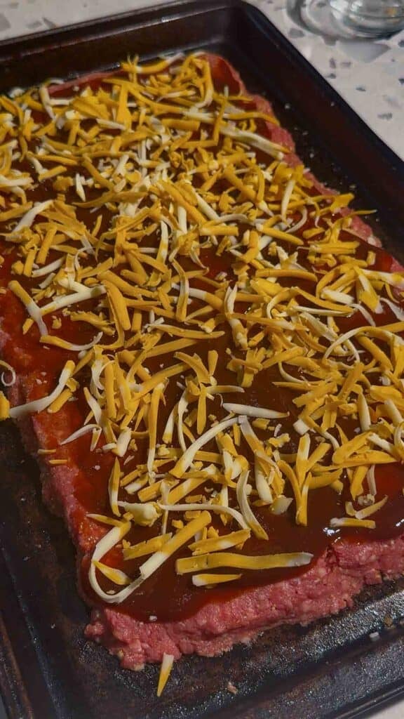 shredded cheddar cheese and sauce mixture on top of raw meatloaf on a sheet pan